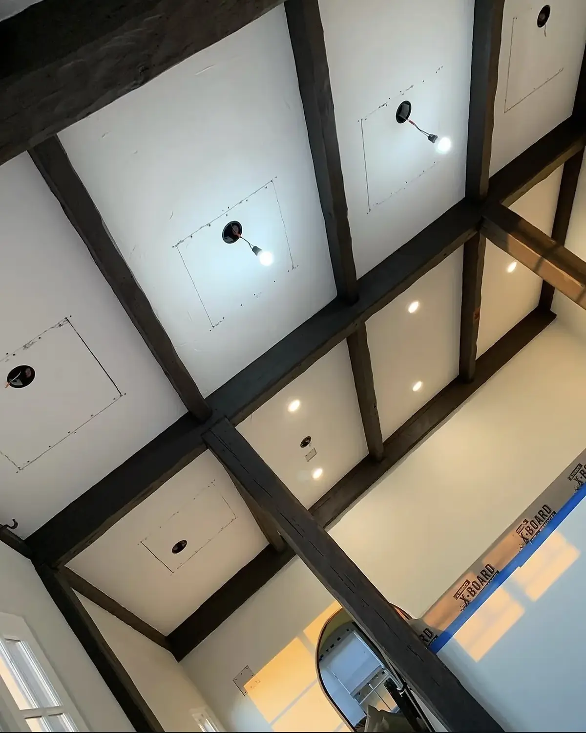 Coffered ceiling with dark beams and white panels, pot light positions outlined in pencil and two openings cut with temporary lamp holders hanging