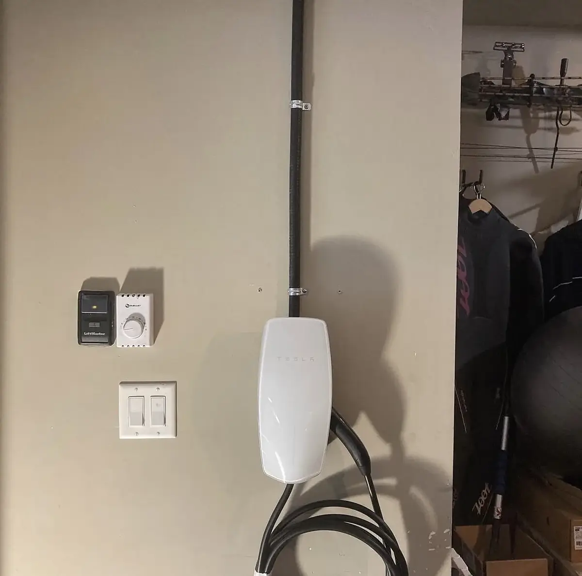 White wall-mounted EV connector on a beige garage wall with conduit running up to the ceiling, a timer switch and light switch beside it and the charging cable coiled below